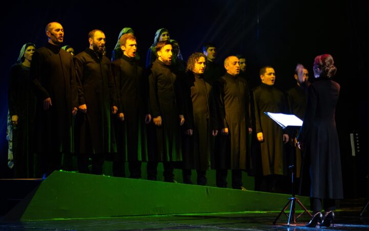 Хор в тёмных сценических костюмах поёт на сцене, на переднем плане дирижёр с нотами