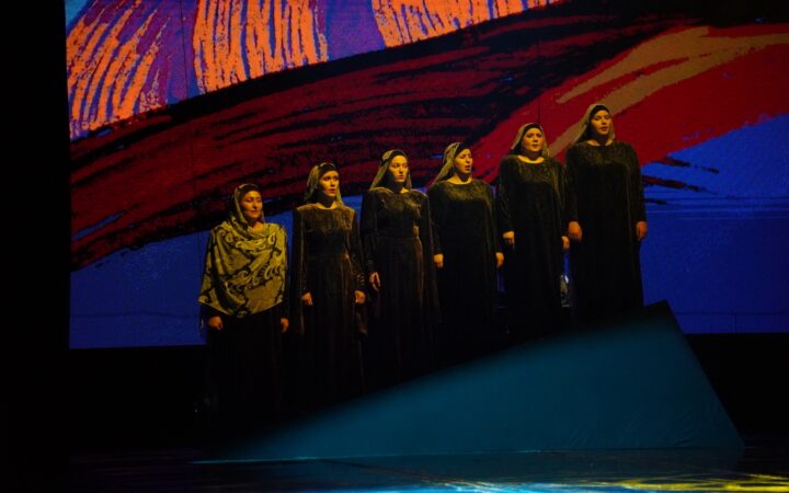 Женский хор в тёмных одеждах на фоне яркой оранжево-синей видеопроекции